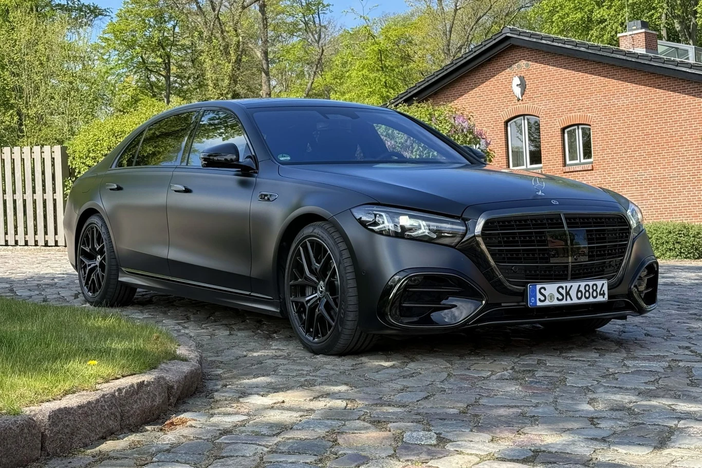 Czarny Mercedes z matowym wykończeniem, sportowymi felgami oraz agresywnym pakietem stylistycznym, zaparkowany na brukowanej drodze przed ceglaną willą.