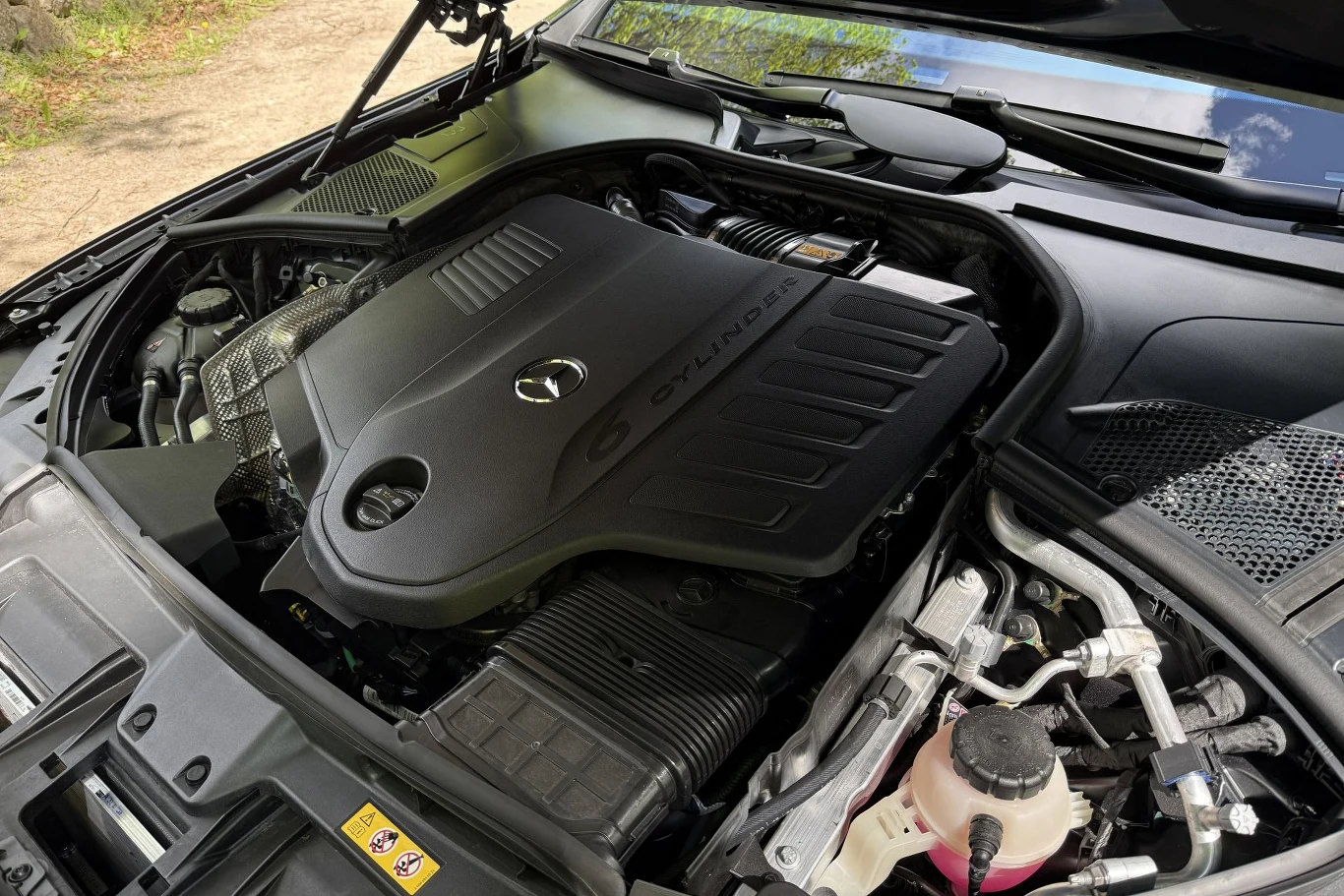 Silnik samochodowy z logo marki Mercedes-Benz z wyraźnie widoczną osłoną silnika oznaczoną napisem „6 CYLINDER”, otoczoną elementami układu chłodzenia i zbiornikiem płynu eksploatacyjnego.