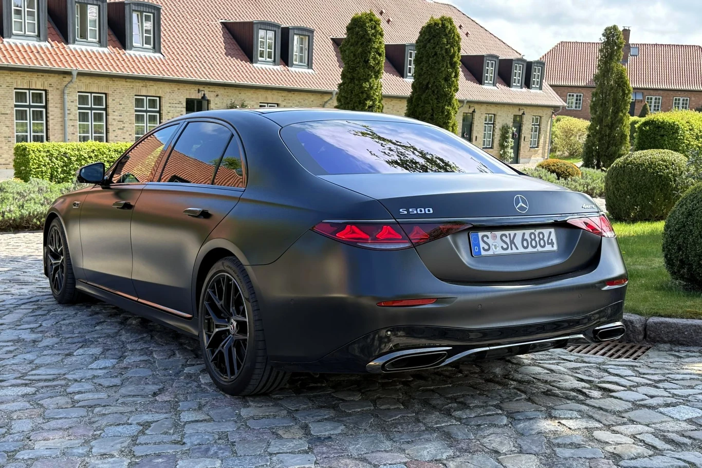 Czarny Mercedes S 500 z przyciemnianymi szybami stoi na brukowanym podjeździe, otoczony elegancką zielenią i zadbanymi krzewami.