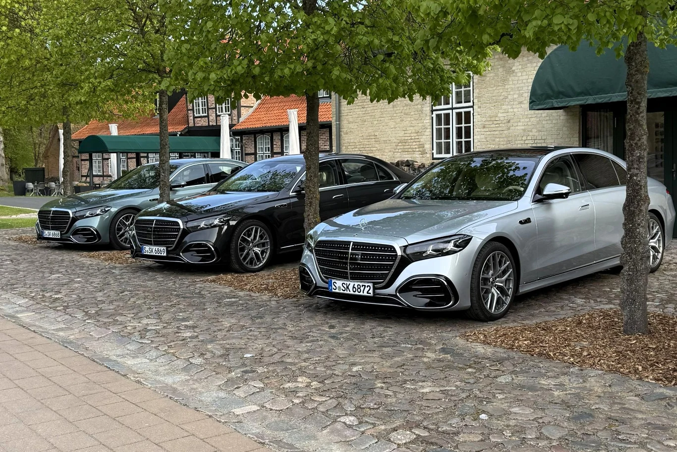 Trzy luksusowe limuzyny Mercedes-Benz zaparkowane obok siebie pod zielonymi drzewami na brukowanym parkingu, na tle kamiennego budynku z czerwonymi dachami.
