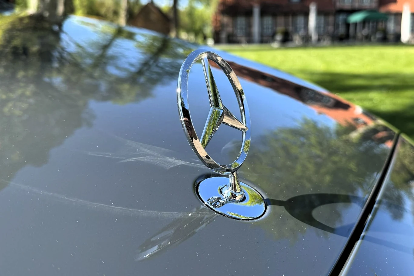 Chromowany emblemat marki Mercedes-Benz osadzony na masce samochodu lśniącej w słońcu.