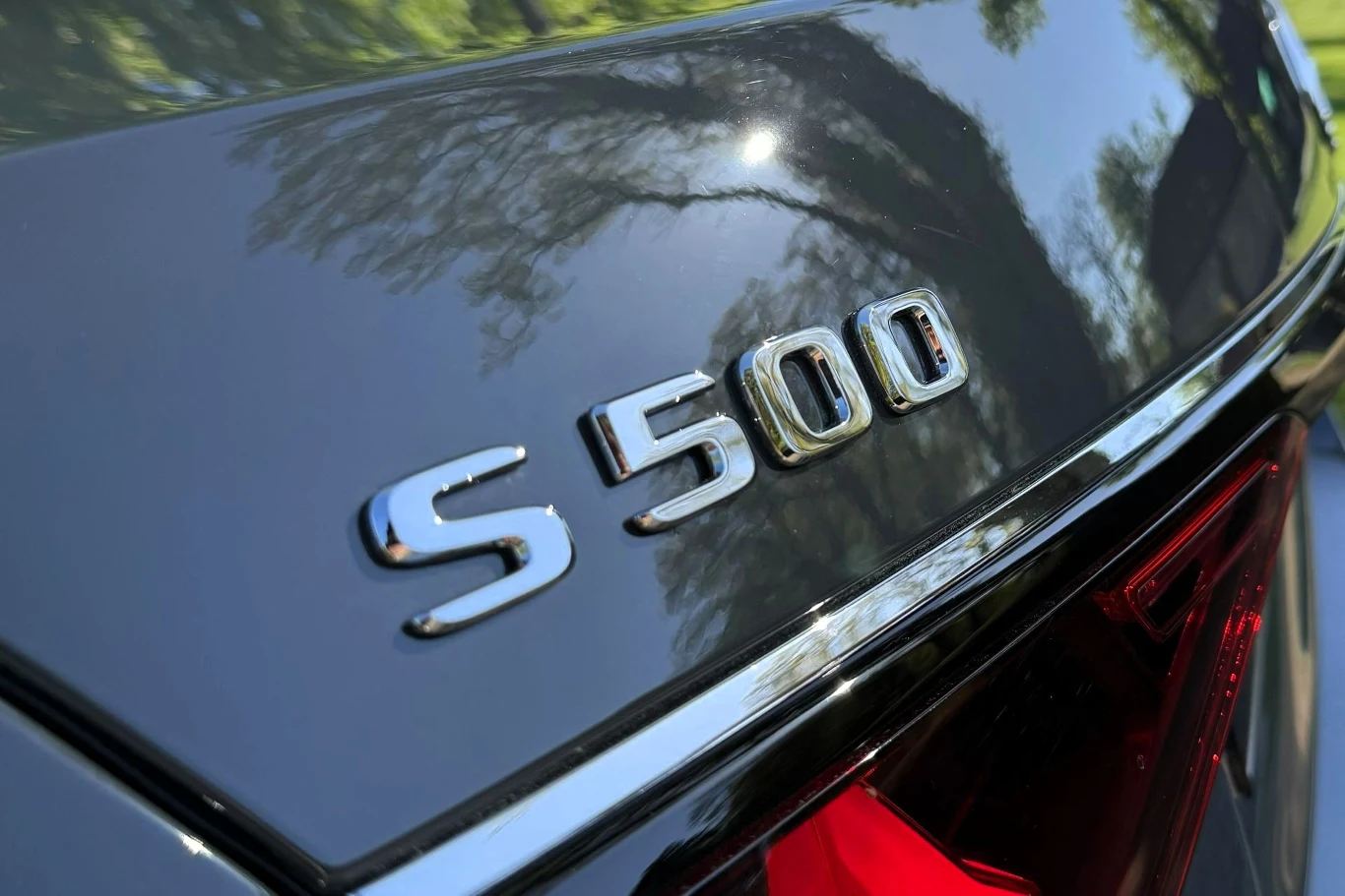 Chromowany emblemat S 500 na tylnej klapie luksusowego auta, obok fragment tylnego światła oraz odbicie drzew na lśniącej karoserii.