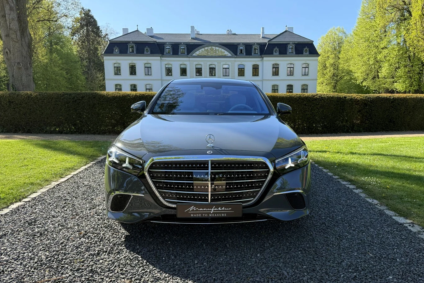 Luksusowy czarny Mercedes klasy S zaparkowany na żwirowej alejce przed eleganckim, białym pałacem otoczonym zielenią.