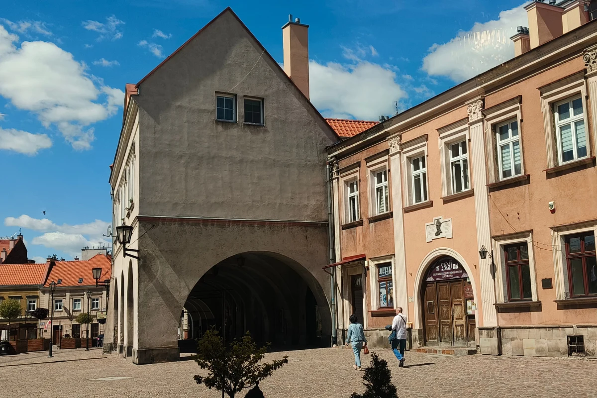 Stare kamienice z jasną fasadą, masywny budynek z dużym łukowatym przejściem oraz dwie osoby spacerujące po brukowanym placu.