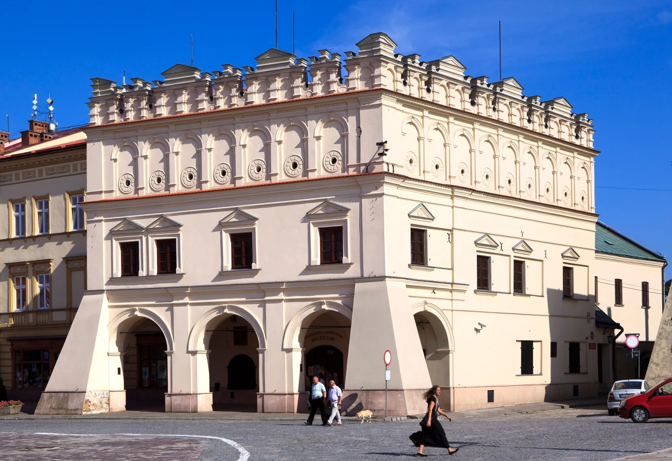 Renesansowa kamienica z charakterystyczną attyką i łukowatymi podcieniami, na tle brukowanego rynku, z grupą przechodniów i psem przechodzącymi przez plac.