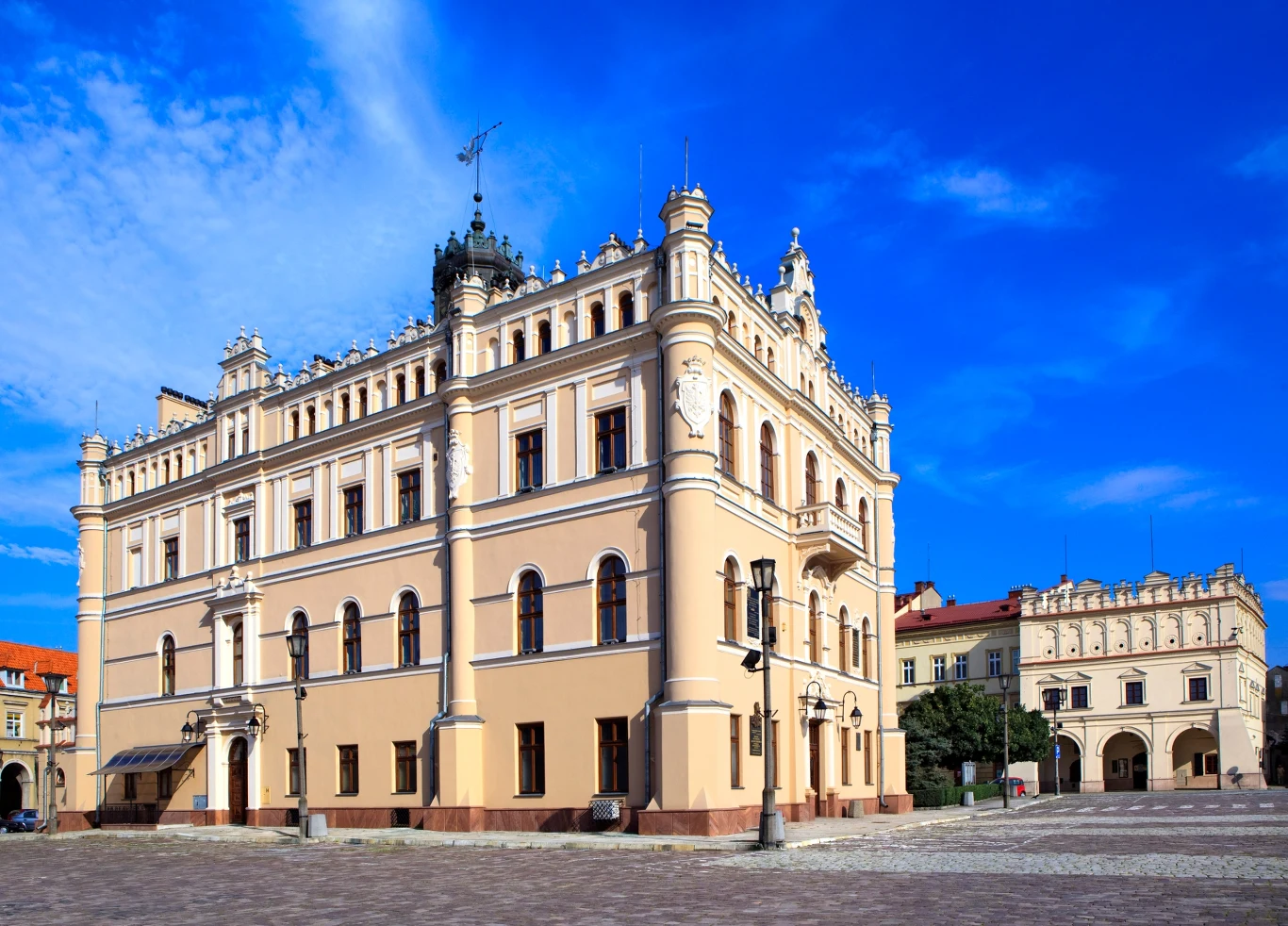 Zabytkowy, jasny budynek o ozdobnej fasadzie z licznymi oknami, detalami architektonicznymi i wieżyczkami, stojący na przestronnym, brukowanym placu pod błękitnym niebem.