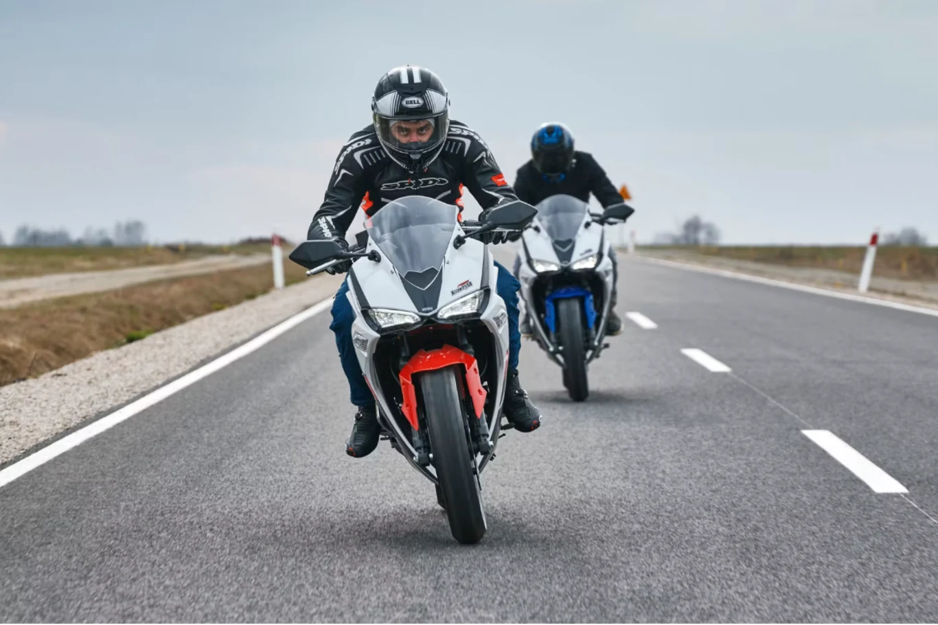 Dwóch motocyklistów w kaskach i kurtkach motocyklowych jedzie sportowymi motocyklami po pustej, asfaltowej drodze, otoczonej płaskim, otwartym krajobrazem.