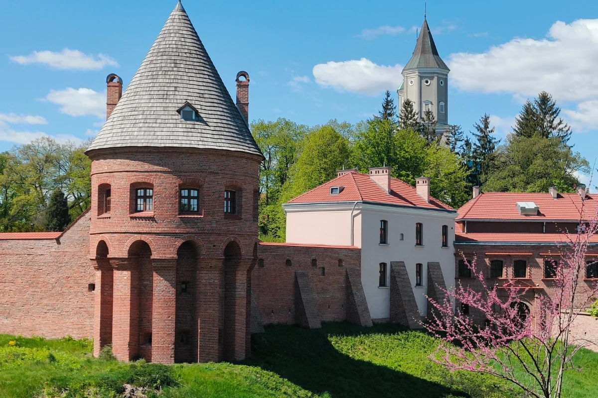 Cylindryczna wieża z cegły z wysokim, stożkowym dachem, otoczona fragmentem murów obronnych oraz przylegającymi do nich zabudowaniami o jasnych elewacjach i czerwonych dachach; w tle na wzgórzu widoczna wieża kościoła otoczona drzewami.