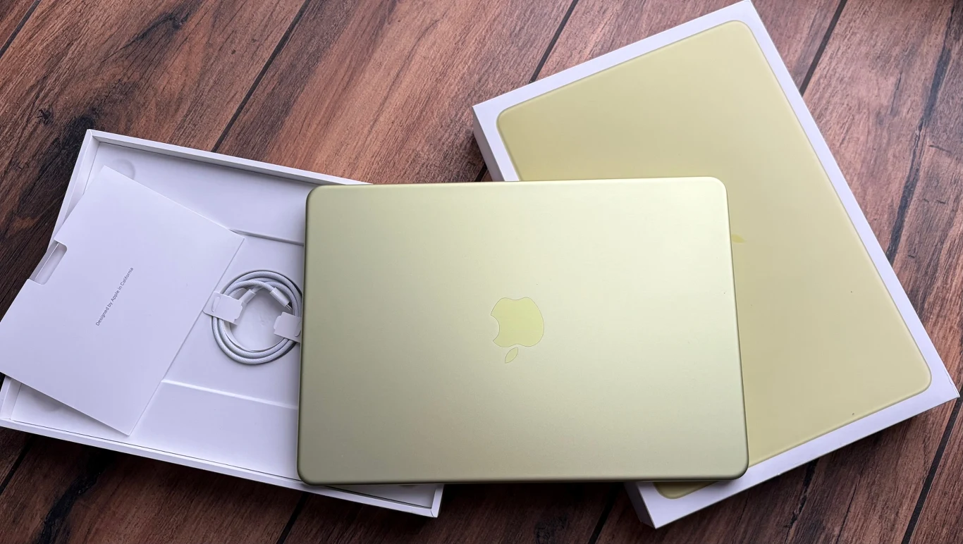 Złoty laptop marki Apple na drewnianej podłodze, częściowo wyjęty z pudełka; widoczna ładowarka i instrukcja obsługi.