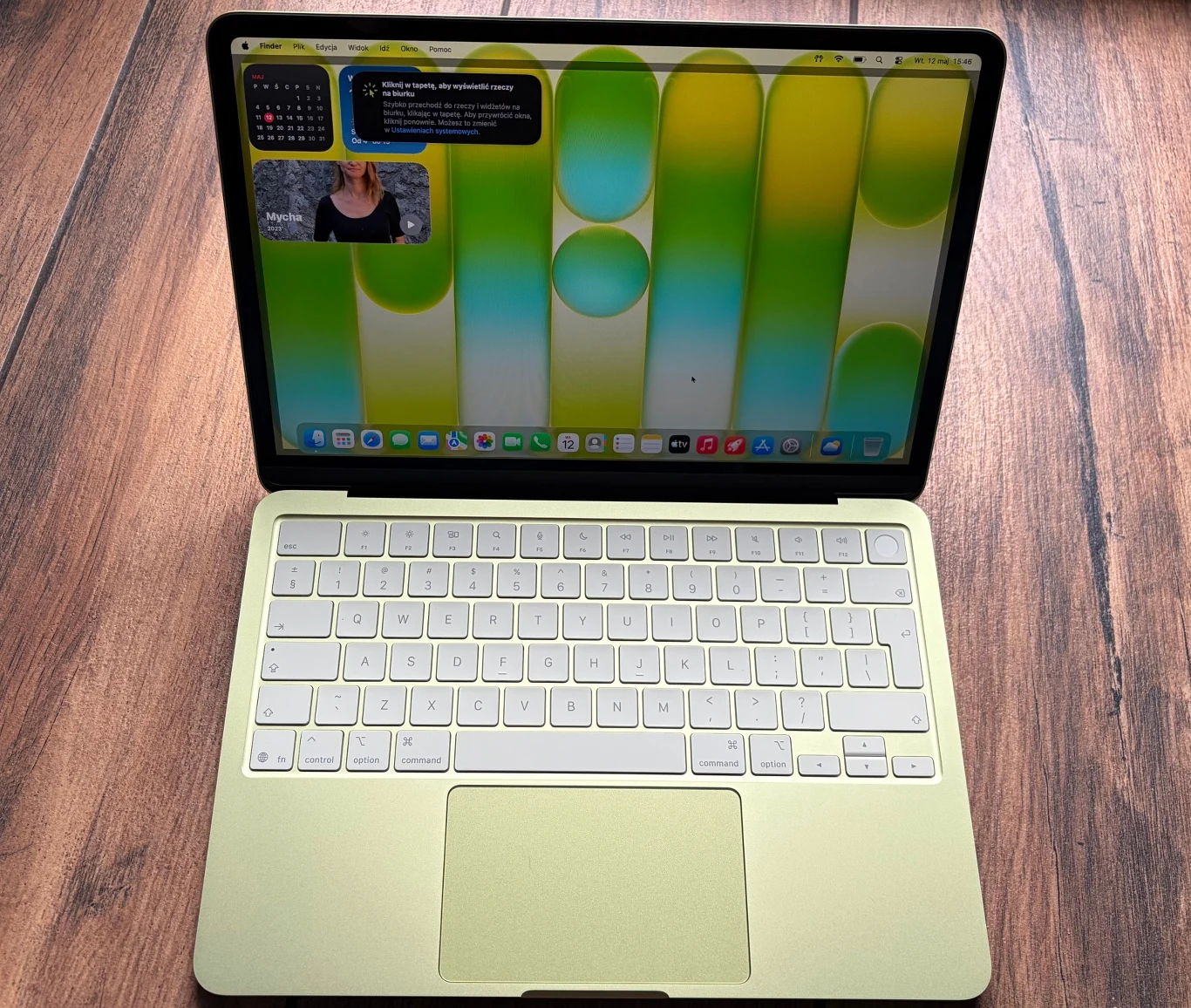 Jasnożółty laptop z dużym wyświetlaczem, na którym widoczny jest kolorowy, abstrakcyjny tapetowy motyw, otwarte aplikacje w lewym górnym rogu oraz minimalistyczna klawiatura z białymi klawiszami.