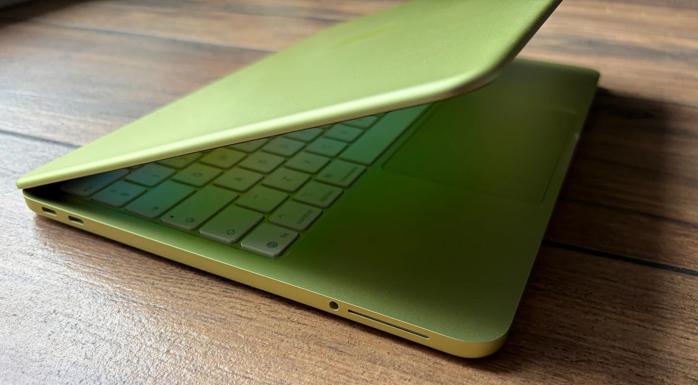 Jasnozielony laptop częściowo otwarty, ustawiony na drewnianej powierzchni, z widoczną białą klawiaturą i smukłą obudową.