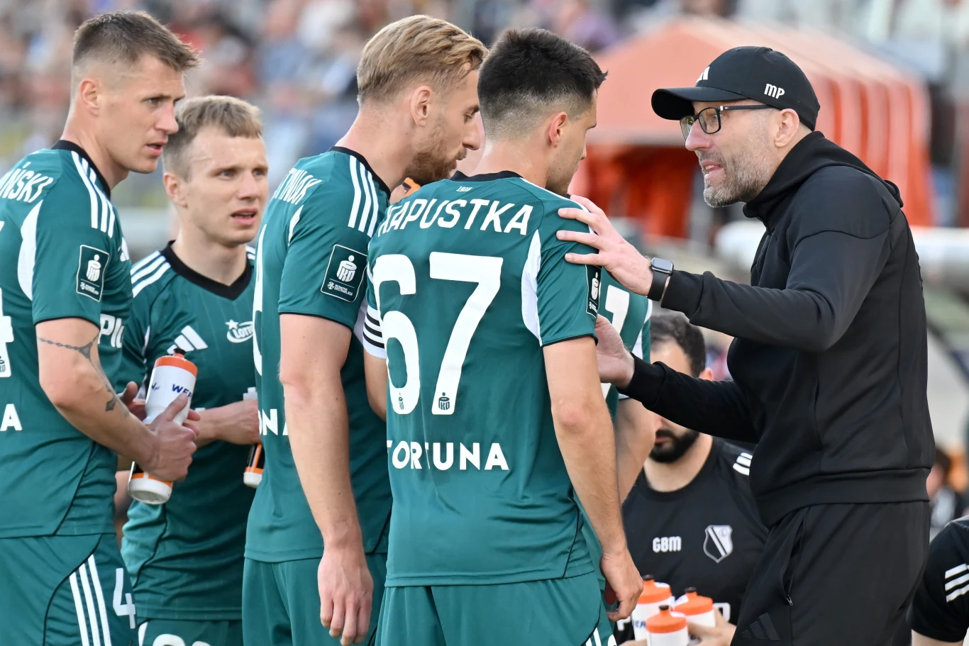 Trener drużyny piłkarskiej w czarnym stroju udziela instrukcji kilku zawodnikom ubranym w zielone koszulki, trzymając jednego z nich za ramię, podczas przerwy w meczu.