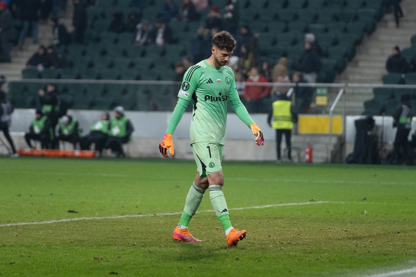 Bramkarz w jasnozielonym stroju z opuszczoną głową opuszcza boisko na stadionie piłkarskim podczas meczu, w tle widoczni są sędziowie techniczni oraz kibice na trybunach.