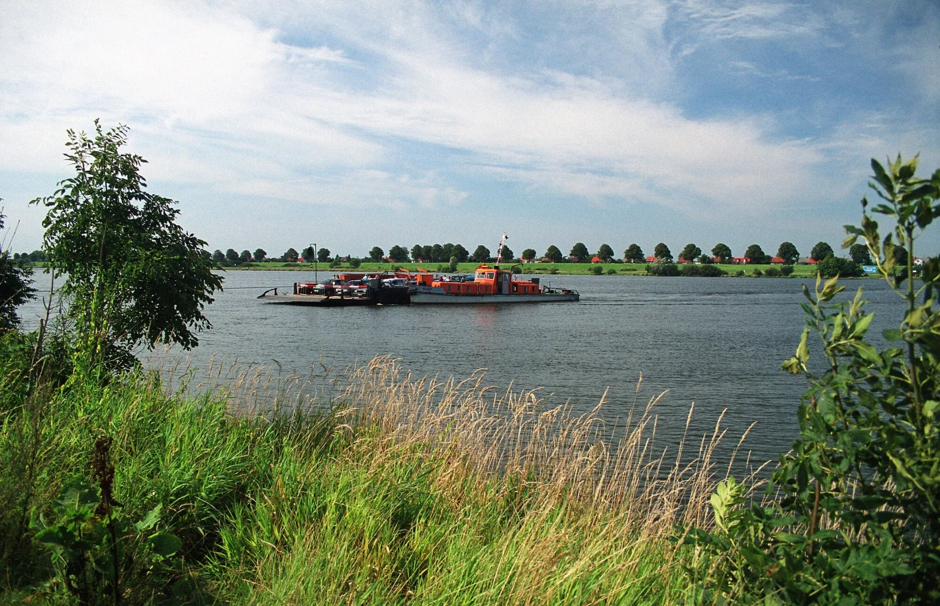 Przeprawa promowa na Wiśle Mikoszewo-Świbno Prom rzeczny z pomarańczowym nadwoziem transportuje pojazdy i ludzi przez szeroką rzekę, w tle równy szpaler drzew na zielonym brzegu.