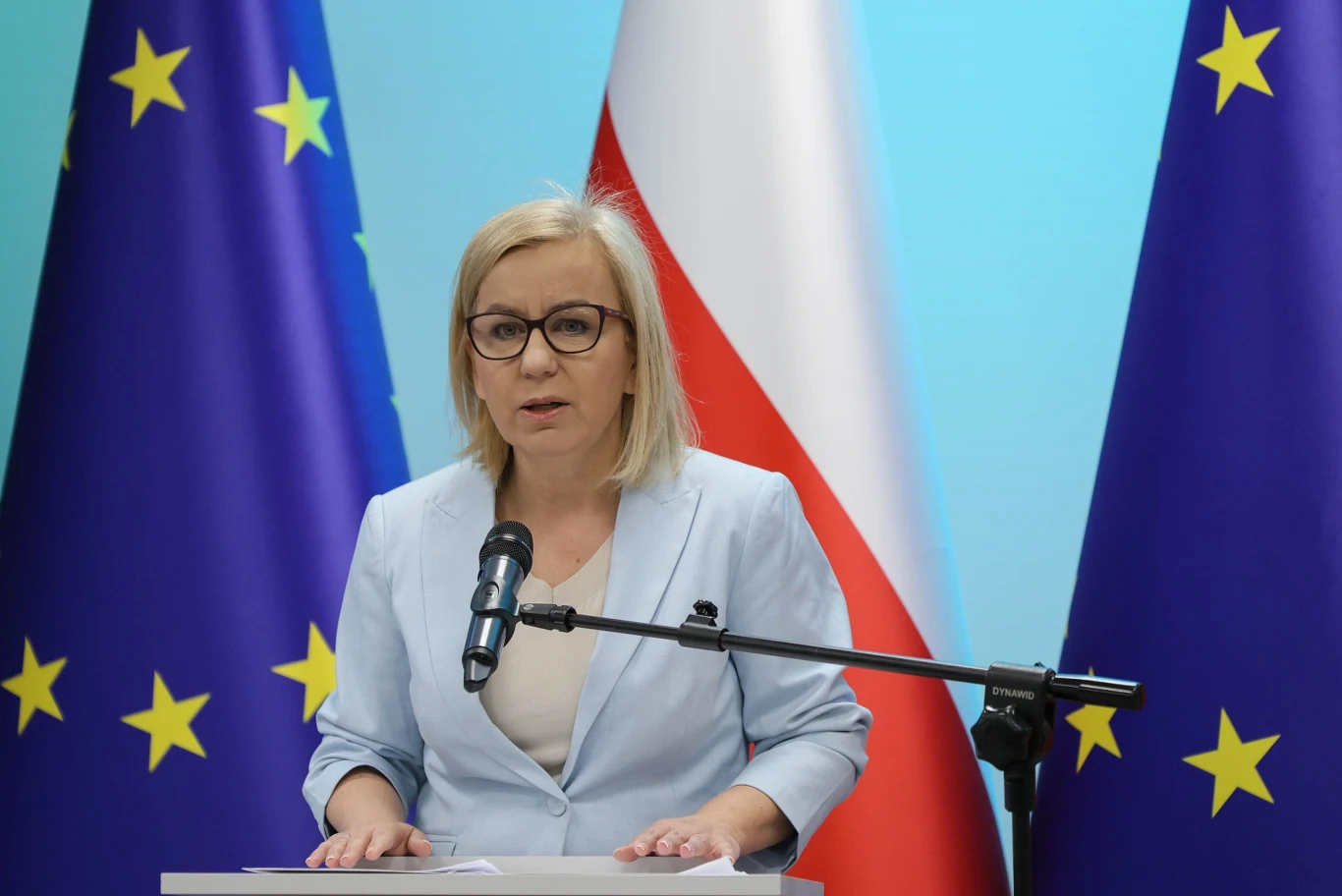 Ogłoszono szczegóły dot. finansowania hubu elektromobilności ze środków KPO. Paulina Hennig-Kloska w jasnym żakiecie przemawia do mikrofonu, w tle flagi UE i Polski.