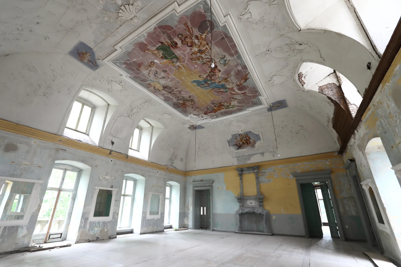 Przestronne, zniszczone wnętrze dawnej sali z dużymi oknami, zdobionym sufitem z barokowym freskiem i kominkiem na ścianie.