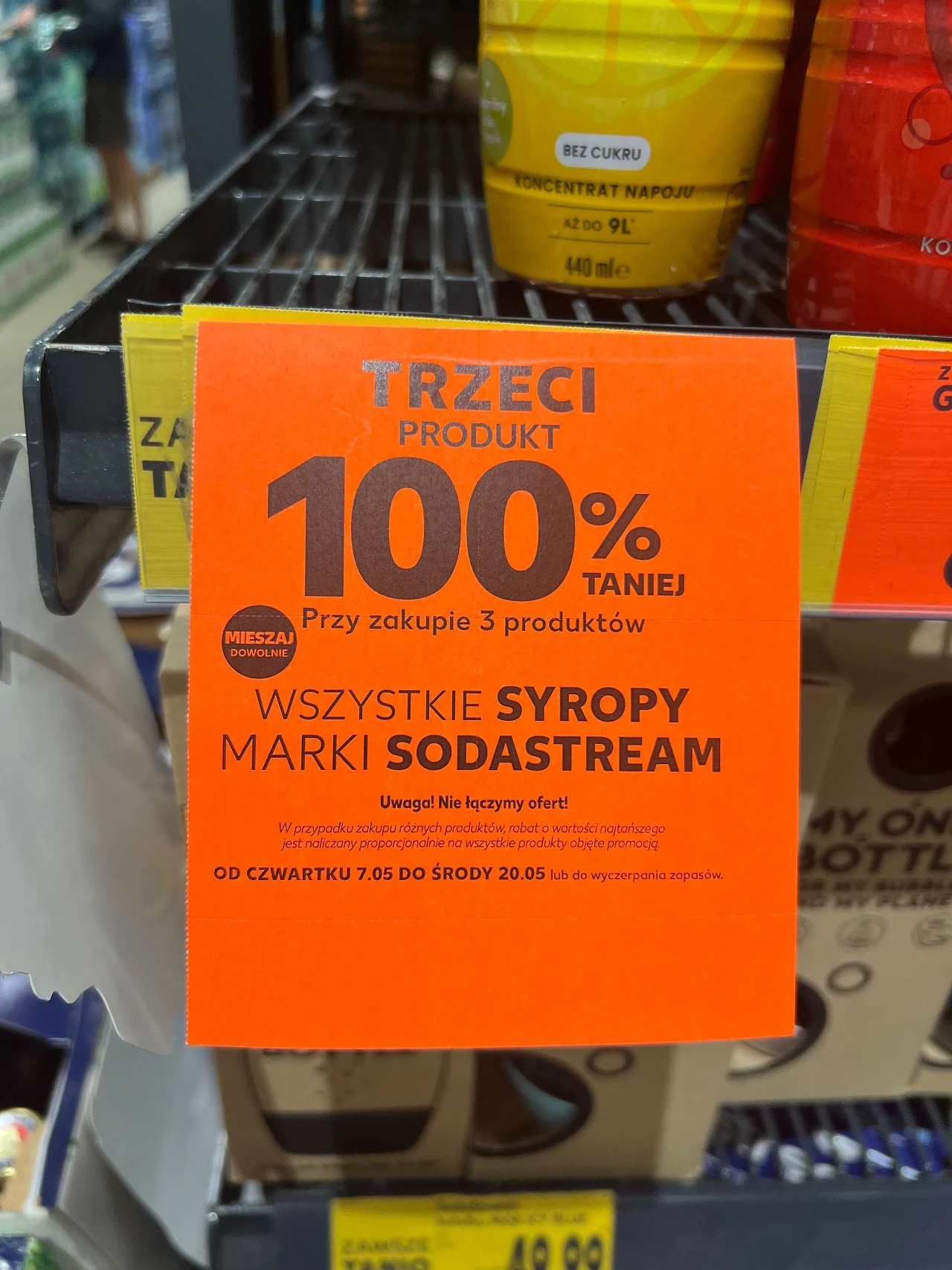 Promocja na syropy Sodastream w Kauflandzie Pomarańczowa etykieta promocyjna informująca o rabacie na syropy marki Sodastream, z napisem o 100% zniżce na trzeci produkt przy zakupie trzech produktów oraz warunkami oferty umieszczonymi na dole plakietki.