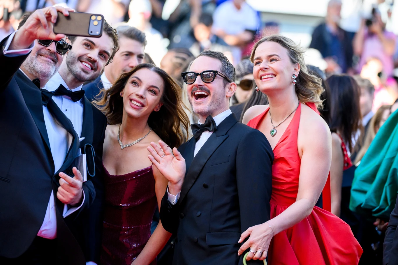 Grupa elegancko ubranych osób uśmiecha się i pozuje do wspólnego selfie podczas oficjalnego wydarzenia, otoczona przez tłum w tle.
