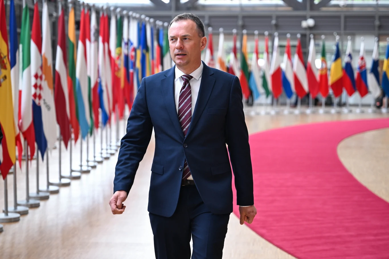 Mężczyzna w garniturze idący po czerwonym dywanie wśród ustawionych w rzędach flag państwowych.