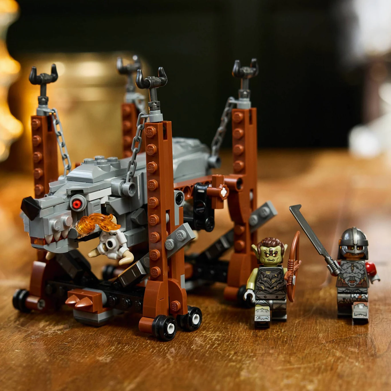 "LEGO Icons: Władca Pierścieni: Grond" Duży, groźny pies z klocków LEGO w otoczeniu dwóch figurek rycerza i potwora — całość utrzymana jest w klimacie fantasy, a zwierzę przykute jest łańcuchami do specjalnej konstrukcji.