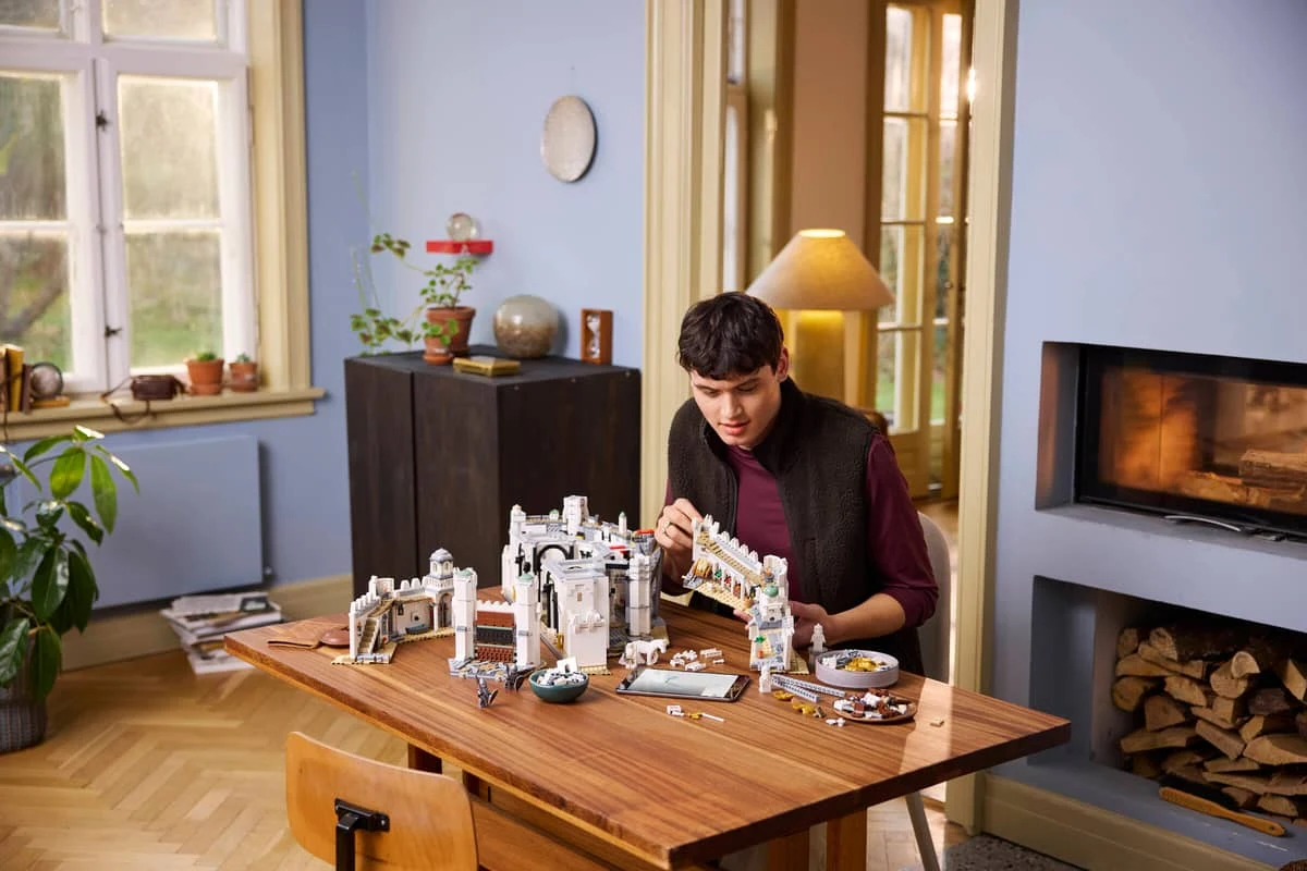 "LEGO Icons: Władca Pierścieni: Minas Tirith" Młody mężczyzna skupiony na składaniu dużego zestawu klocków LEGO na drewnianym stole w jasnym, przytulnym salonie z kominkiem i nowoczesnym wystrojem.