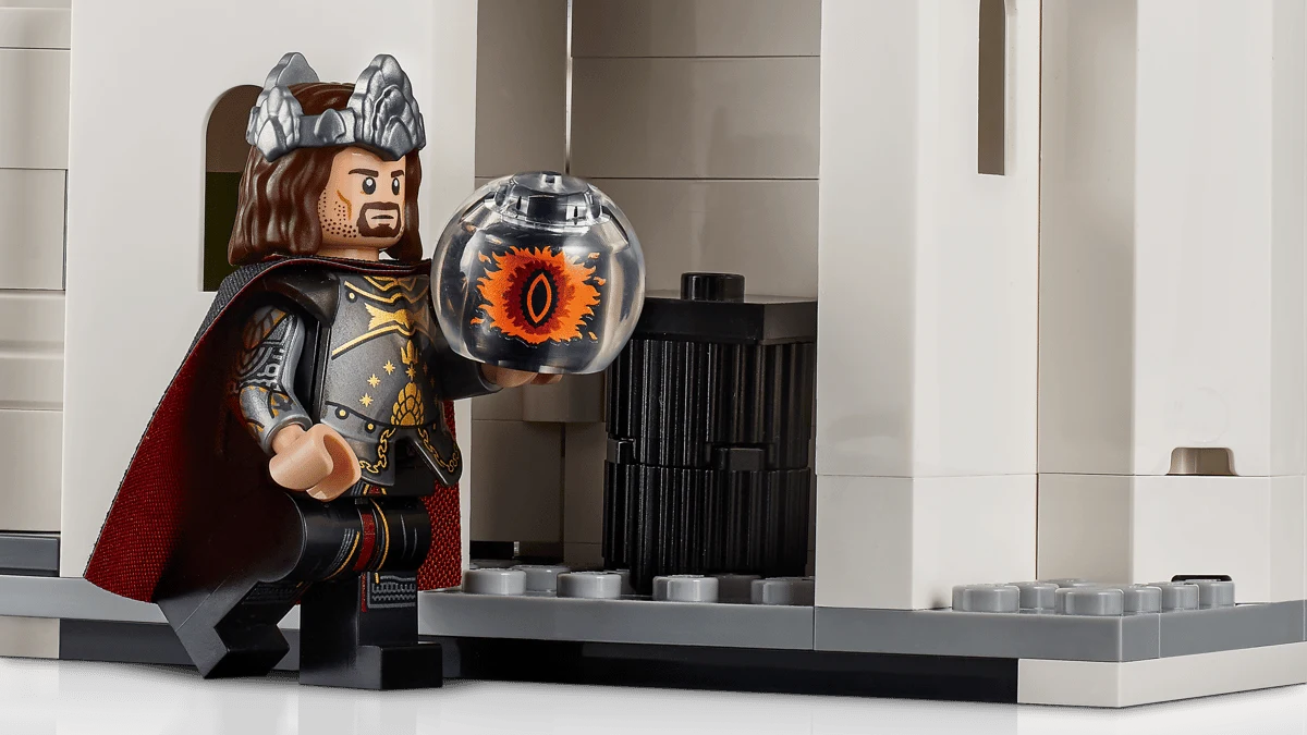 "LEGO Icons: Władca Pierścieni: Minas Tirith" Figurka LEGO w stroju rycerza z peleryną i hełmem trzyma kulę z symbolem oka, stojąc obok jasnej budowli z klocków.