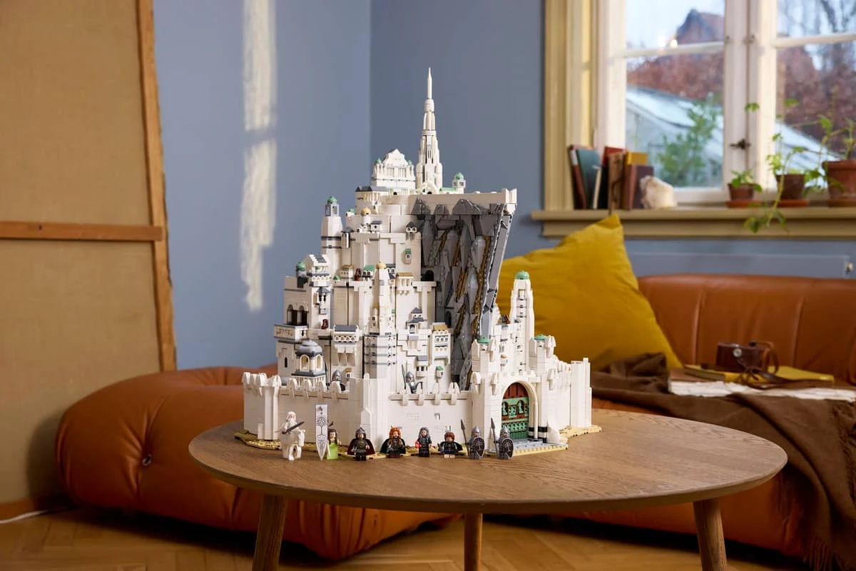 "LEGO Icons: Władca Pierścieni: Minas Tirith" Duży model białego zamku z klocków Lego ustawiony na okrągłym drewnianym stoliku, przed którym stoi rząd figurek postaci z serii Władca Pierścieni; w tle jasne poduszki na brązowej sofie i kilka roślin na parapecie okna.
