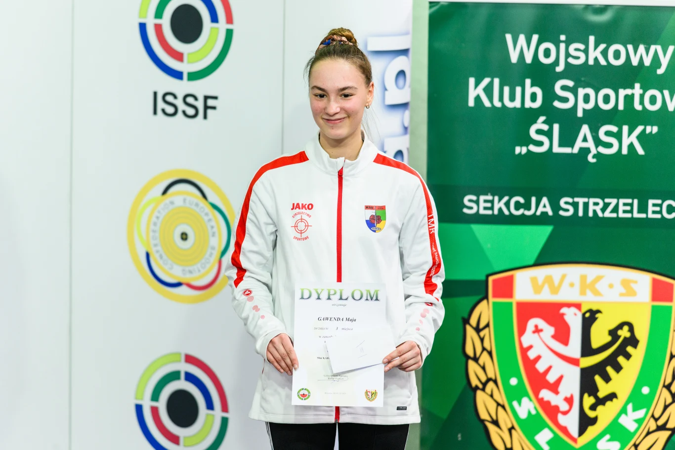 Maja Gawenda Młoda kobieta w sportowej białej bluzie z herbem na piersi trzymająca dyplom, stojąca na tle zielonego banera Wojskowego Klubu Sportowego „Śląsk” i tablicy z kolorowymi emblematami strzeleckimi.