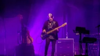 Gitarzysta grający na gitarze basowej na scenie oświetlonej fioletowym światłem, w otoczeniu sprzętu muzycznego i instrumentów.