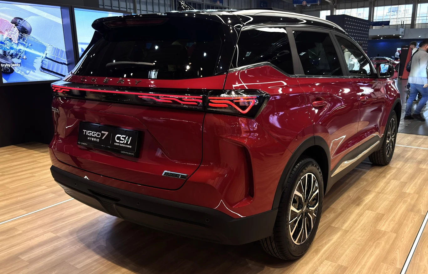Ceny modelu TIGGO 7 Hybrid (HEV) startują od 124 900 złotych Czerwony SUV Tiggo 7 z przyciemnionymi szybami, czarnymi felgami i nowoczesnym, poziomym pasem świateł z tyłu, ustawiony na ekspozycji targowej z drewnianą podłogą.