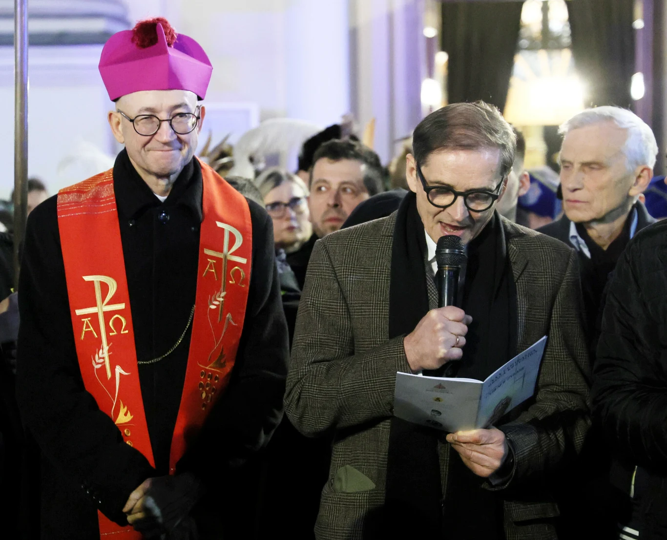 Biskup w fioletowej piusce i czerwonym ornacie stoi obok mężczyzny czytającego do mikrofonu przy tłumie zgromadzonych ludzi.