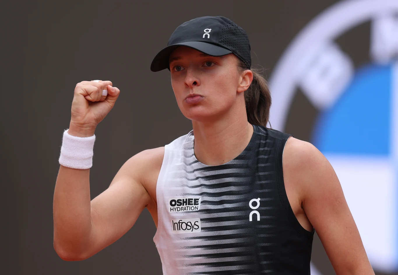 Tenisistka w sportowym stroju i czapce, z uniesioną w geście triumfu zaciśniętą pięścią oraz skupionym wyrazem twarzy.