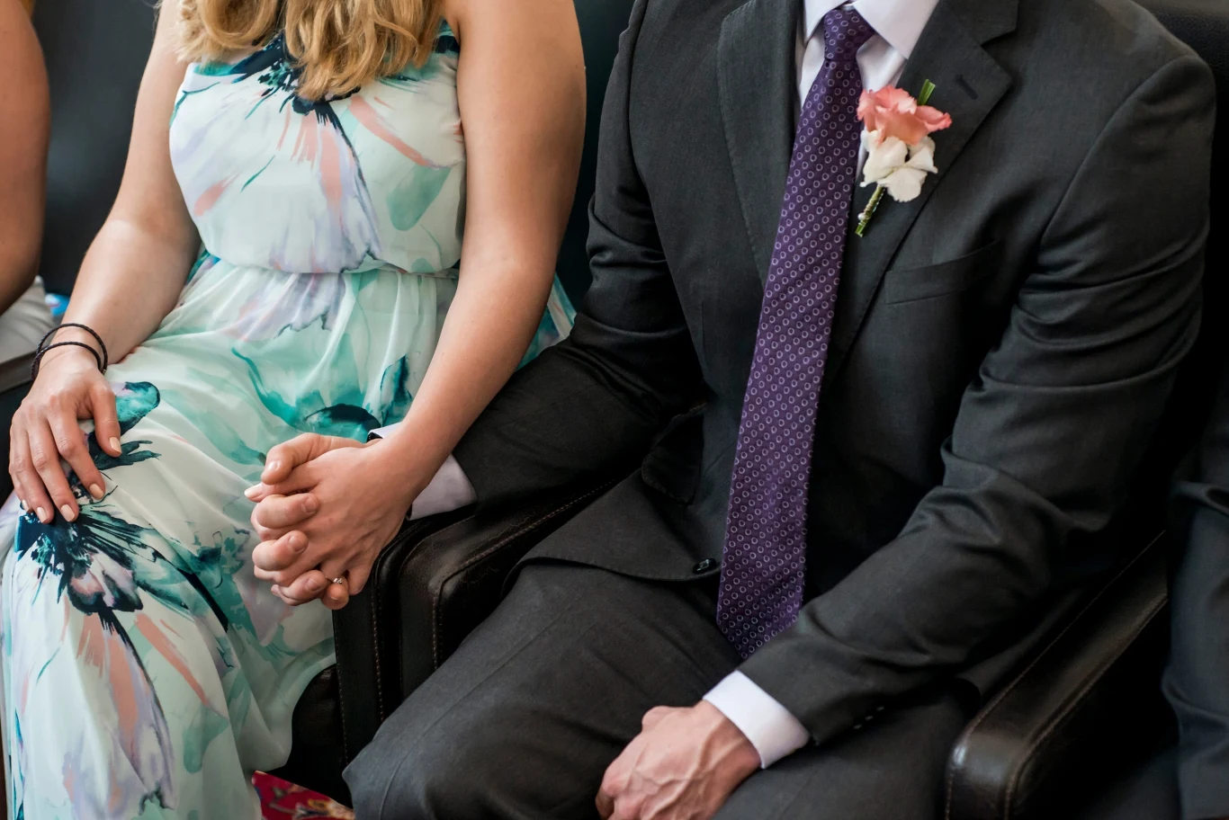 Para elegancko ubranych osób siedzi razem, trzymając się za ręce; kobieta ma na sobie jasną sukienkę w kwiaty, a mężczyzna ciemny garnitur, białą koszulę, fioletowy krawat i butonierkę z kwiatem.