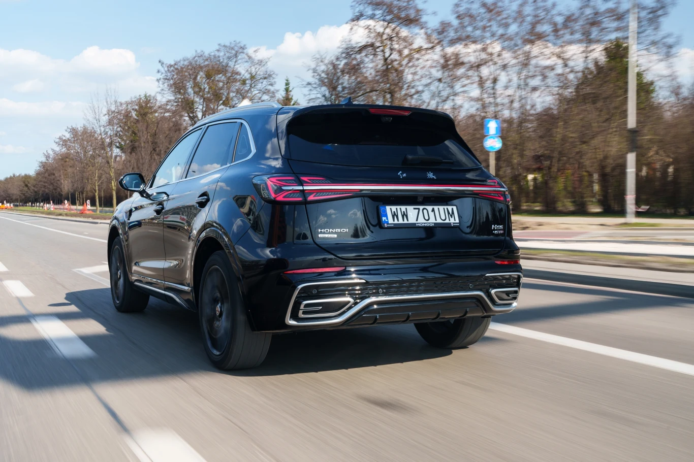 Czarny SUV marki Renault jedzie miejską ulicą, widoczny z tyłu w ruchu, otoczony drzewami bez liści i niską zabudową po prawej stronie.