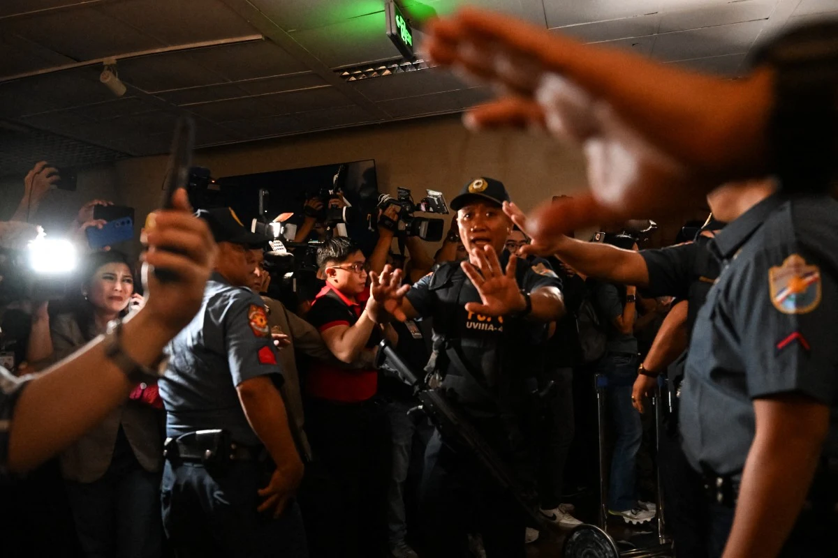 Policjanci w mundurach odgradzają tłum dziennikarzy i fotoreporterów, podkreślając napiętą atmosferę sytuacji.