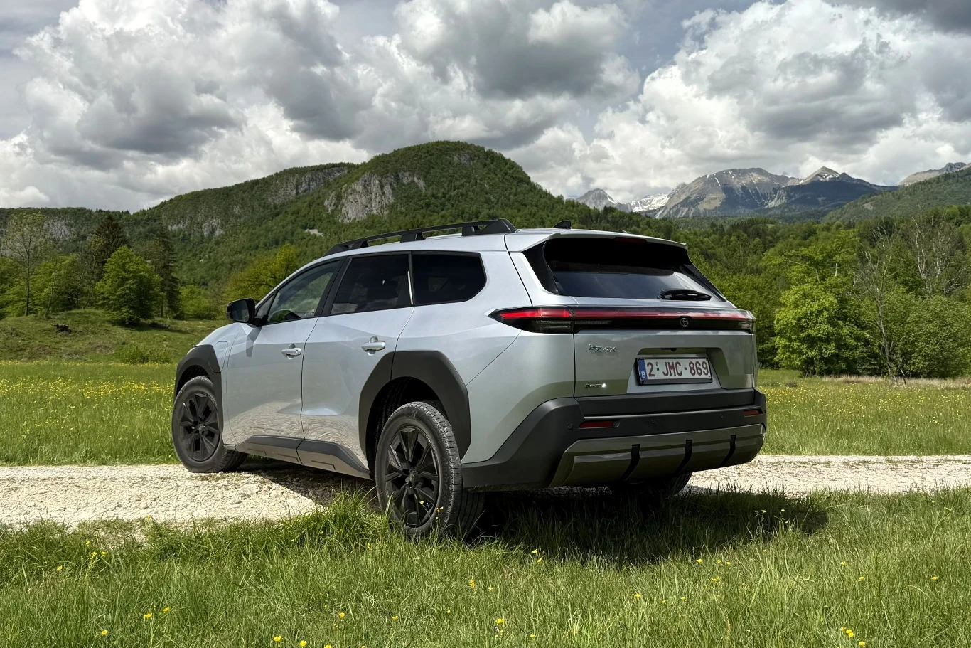 Srebrny SUV zaparkowany na szutrowej drodze pośród zielonych łąk, w tle masywne, zalesione wzgórza i zaśnieżone szczyty gór pod zachmurzonym niebem.