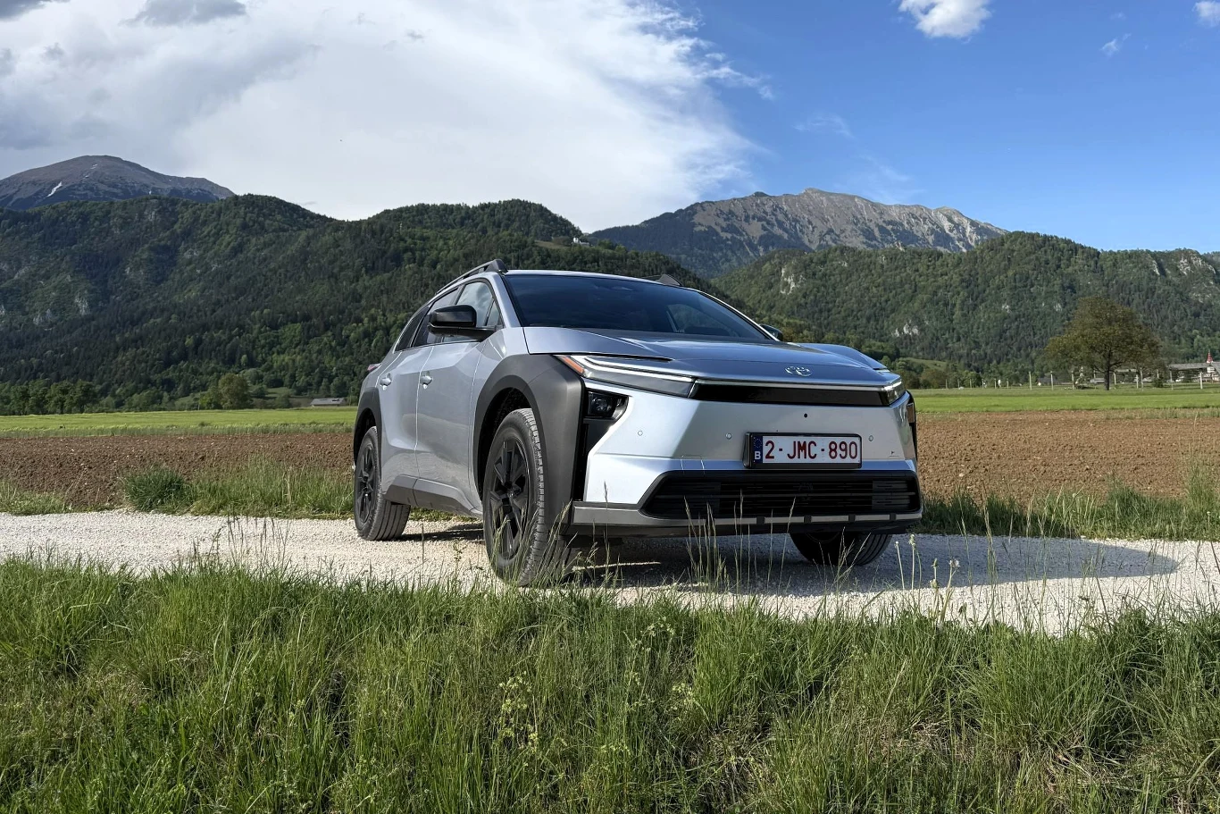 Nowoczesny srebrny SUV zaparkowany na szutrowej drodze z widokiem na zielone pola i górskie wzgórza w tle.