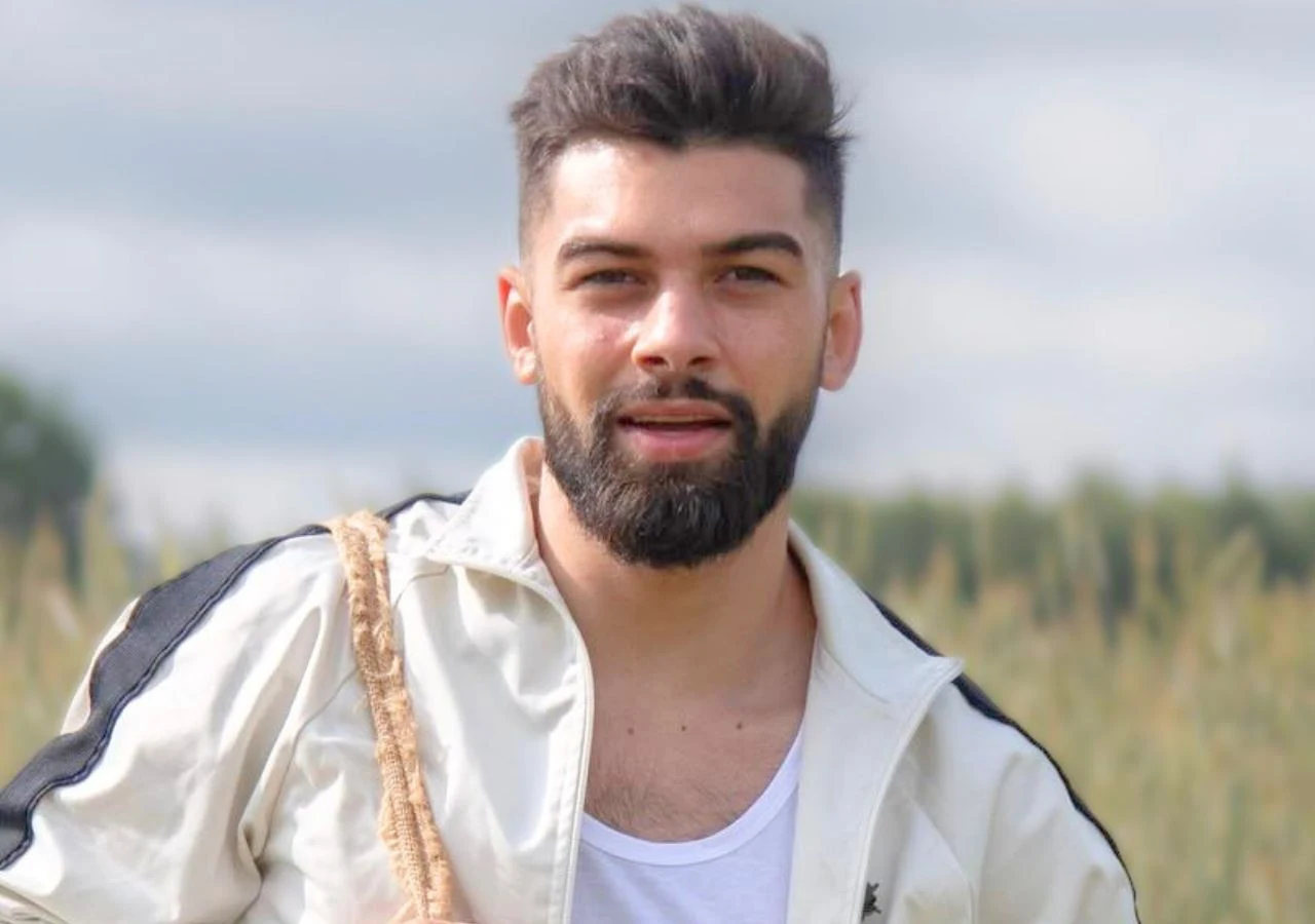 Aksel w programie "Farma" Aksel Rumenov, zwycięzca 5. edycji programu "Farma", stoi na tle wiejskiego krajobrazu w sportowej kurtce.
