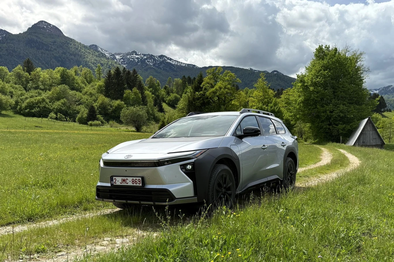 Srebrny samochód typu SUV jedzie po wąskiej, szutrowej drodze otoczonej zielonymi łąkami, w tle widoczne są zalesione wzgórza oraz ośnieżone szczyty gór.