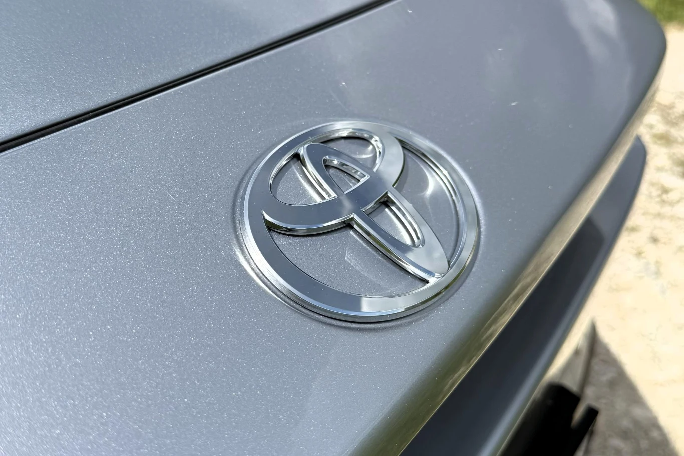 Srebrne logo marki Toyota umieszczone na jasnoszarej karoserii samochodu.
