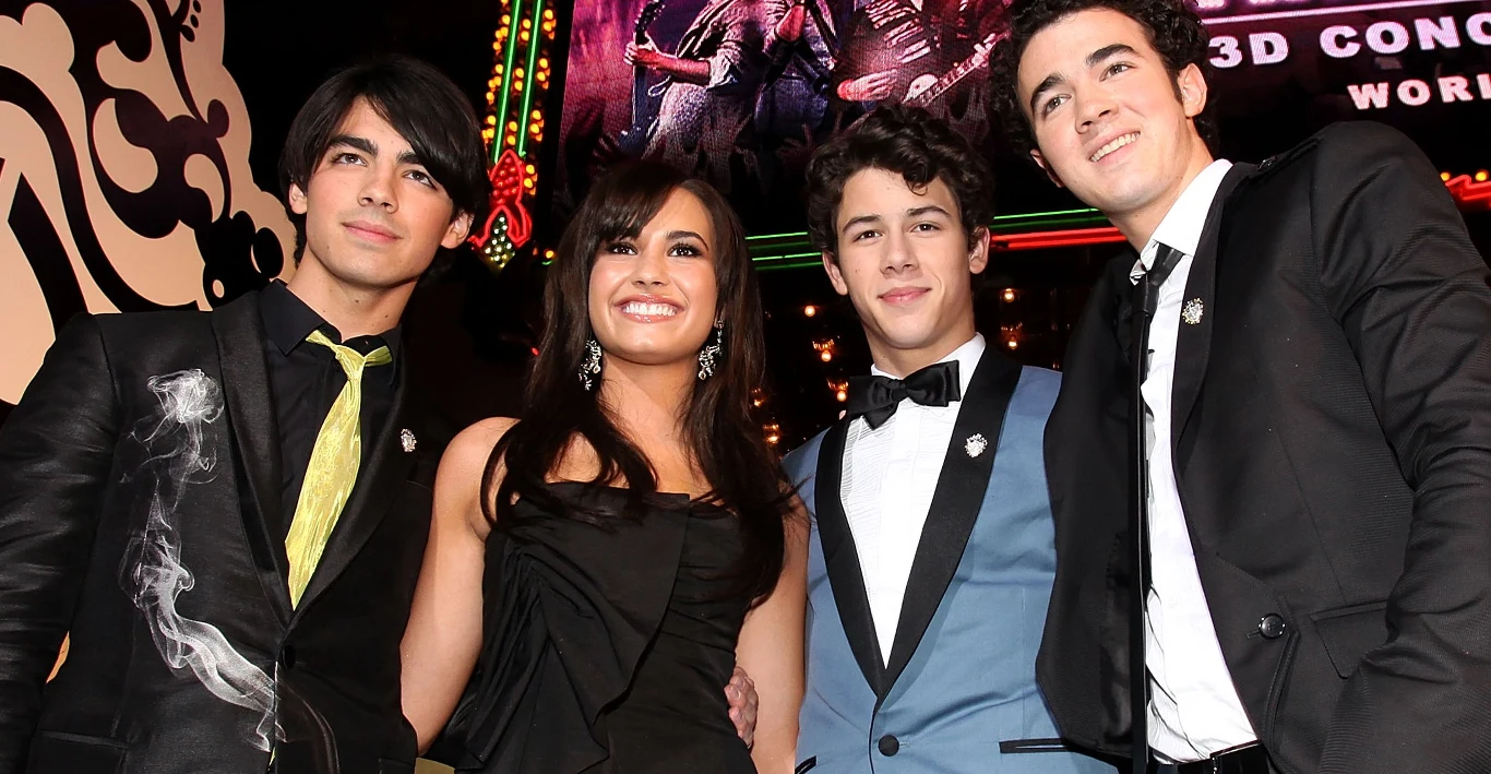 Demi Lovato i Jonas Brothers Demi Lovato i Jonas Brothers w eleganckich strojach wieczorowych na tle świateł i plakatu filmowego