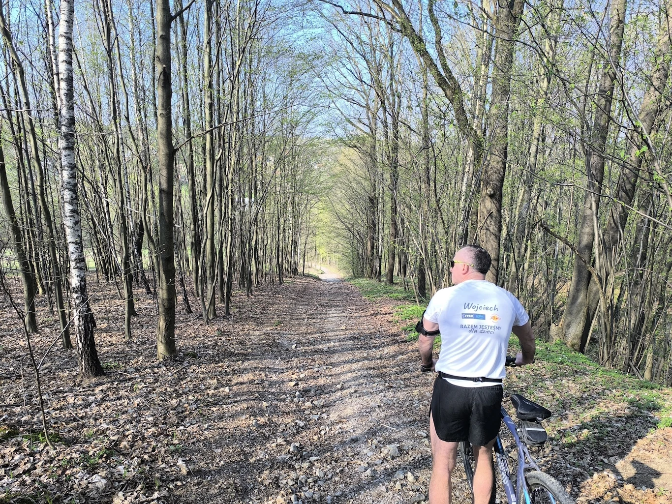 Mężczyzna w sportowym stroju z rowerem stoi na leśnej ścieżce otoczonej smukłymi drzewami, obserwując prostą, piaszczystą drogę prowadzącą w głąb lasu.