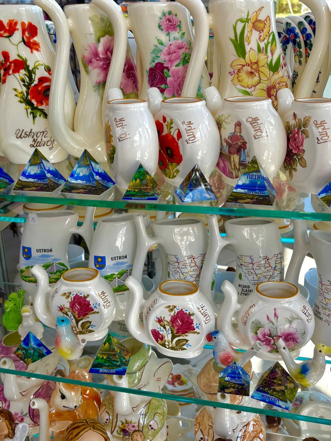 Półki z kolorowymi ceramicznymi dzbankami i filiżankami ozdobionymi kwiatowymi motywami oraz pamiątkowymi miniaturkami szklanych piramid z napisami i nazwami miejscowości.