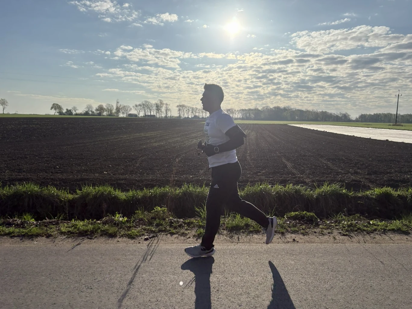Mężczyzna biegnący asfaltową drogą wzdłuż pola uprawnego przy porannym słońcu, otoczony naturalnym krajobrazem.