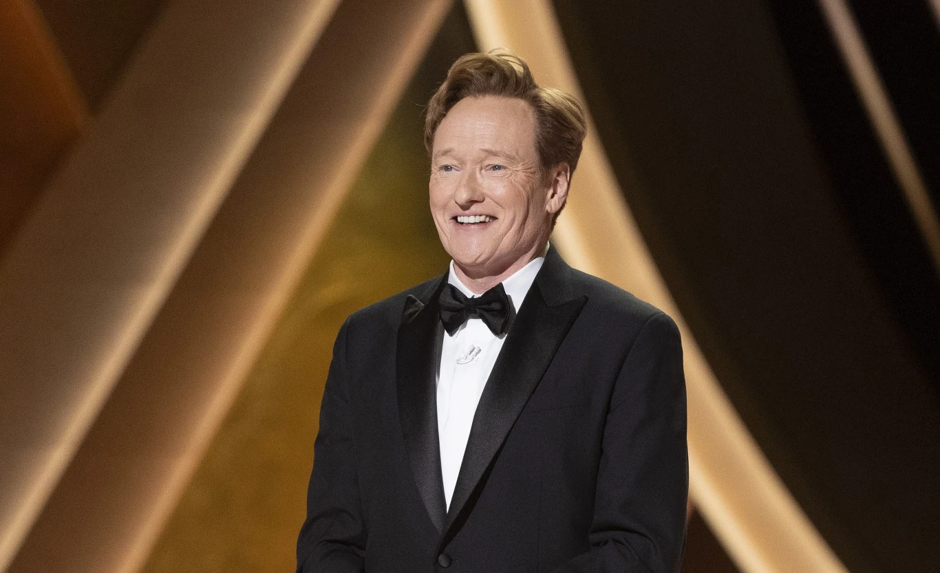 Conan O'Brien Conan O'Brien w smokingu i muszce, na tle geometrycznych dekoracji w ciepłych kolorach.
