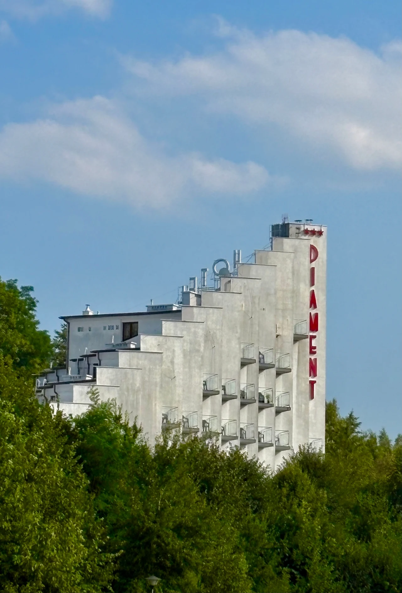 Modernistyczny, biały budynek hotelu Diament o charakterystycznej kaskadowej bryle, wyłaniający się spośród drzew pod błękitnym niebem.