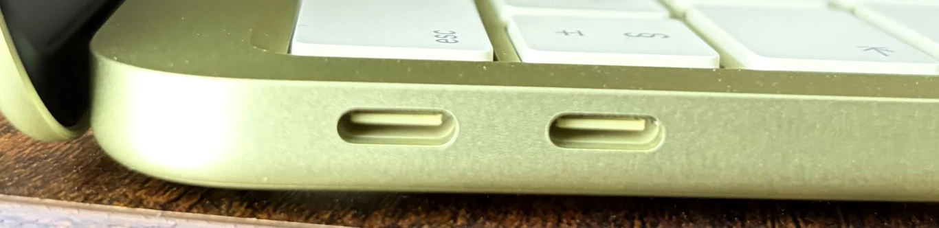 Dwa porty USB-C umieszczone w jasnozielonej obudowie laptopa, fragment białej klawiatury oraz część drewnianej powierzchni, na której stoi urządzenie.