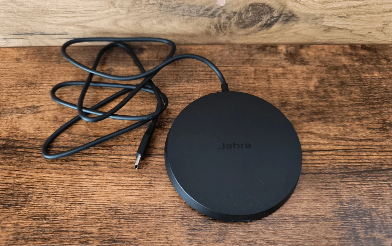 Czarna, okrągła ładowarka Jabra z wyraźnie widocznym przewodem USB-C, umieszczona na drewnianej powierzchni.