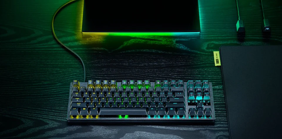 Mechaniczna klawiatura z podświetleniem RGB ustawiona na biurku obok monitora emitującego kolorowe światło.