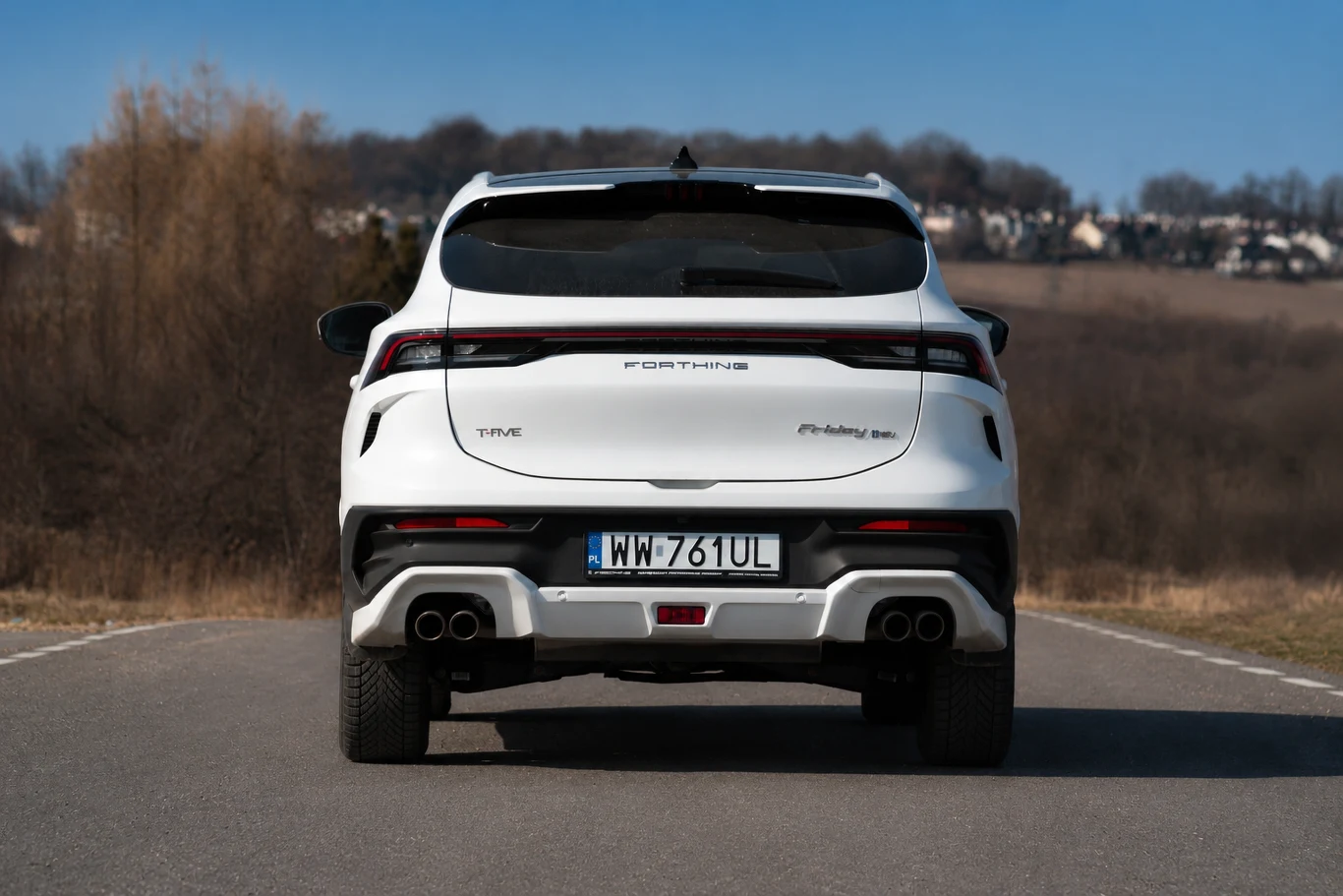Biały samochód typu SUV z widoczną tablicą rejestracyjną, masywnym zderzakiem tylnym i dwoma podwójnymi końcówkami wydechu, ustawiony na wiejskiej drodze z krajobrazem pól i drzew w tle.